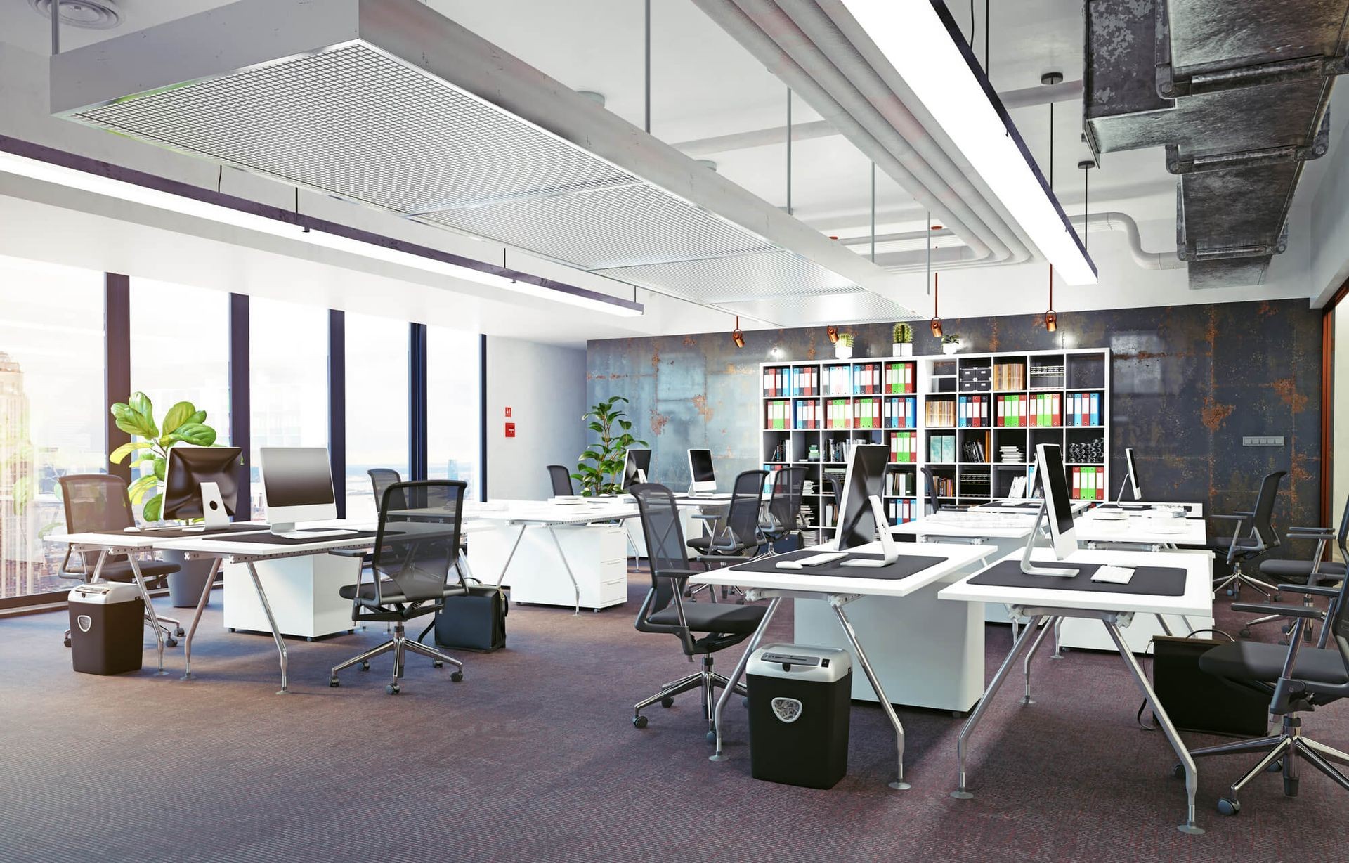 Modern office space with desks, computers, ergonomic chairs, and bookshelves against a large window.